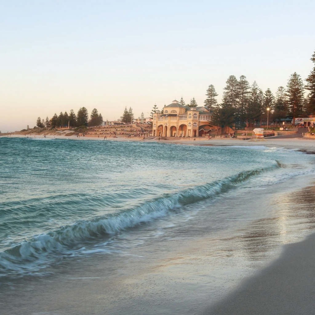 Gentle waves rolling onto the sand at Cottesloe Beach with the historic Indiana Tea House and Norfolk pine trees along the shoreline at sunset in Perth, Western Australia.